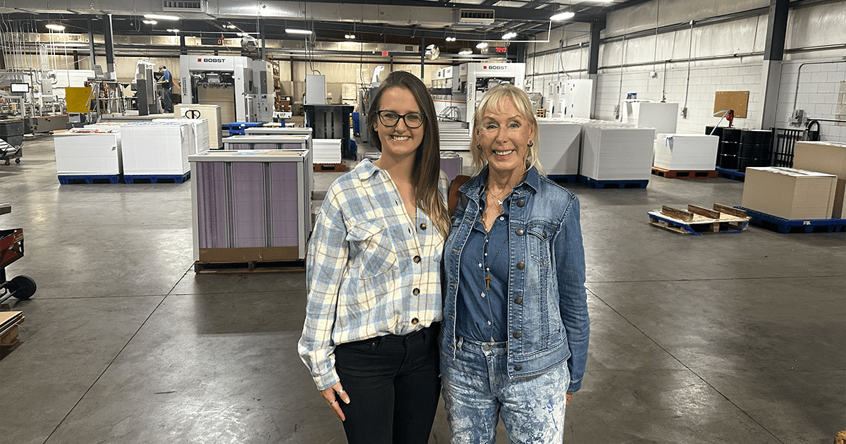 Jan and Hannah smiling in the middle of a large folding carton manufacturing plant, symbolizing the critical role of business and customer service in the skilled trades.