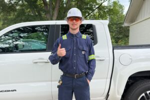 Walter, an industrial electrician, giving a thumbs up in his hard hat, safety glasses, and work uniform in front of a white pickup truck