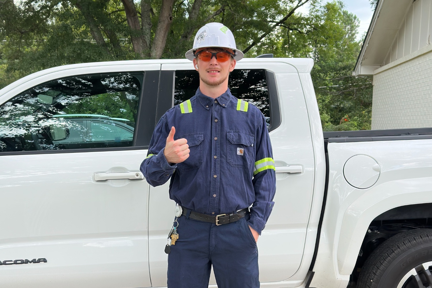 Walter, an industrial electrician, giving a thumbs up in his hard hat, safety glasses, and work uniform in front of a white pickup truck