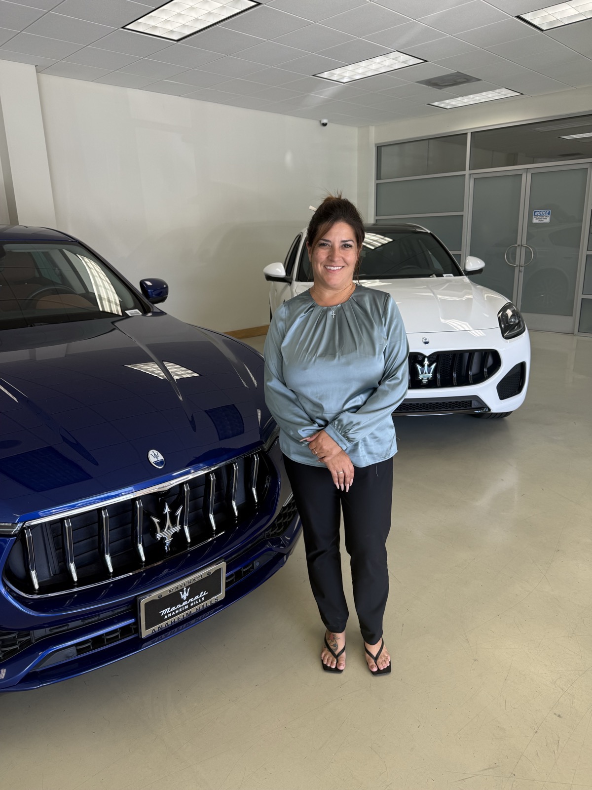 Christina, an automotive service manager, standing between a blue and a white Maserati SUV in the dealership showroom