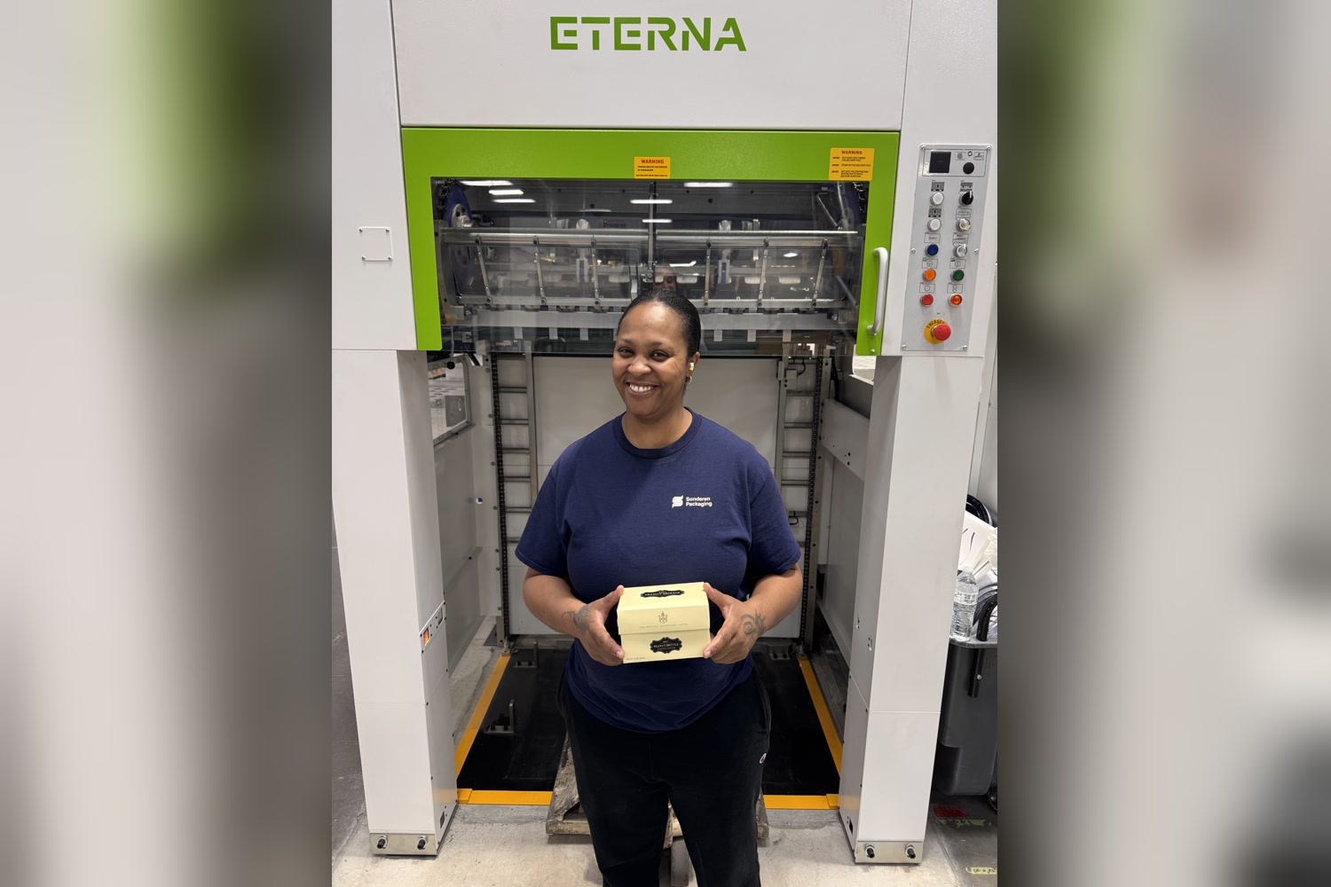 Techelle, a manufacturing cutter and foil operator, holding a foil-stamped box in front of the ETERNA foil stamping machine she runs