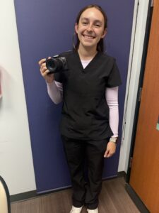 Lauren, a 23-year-old medical photographer, smiling in black scrubs and holding her Canon camera at the dermatology clinic where she works